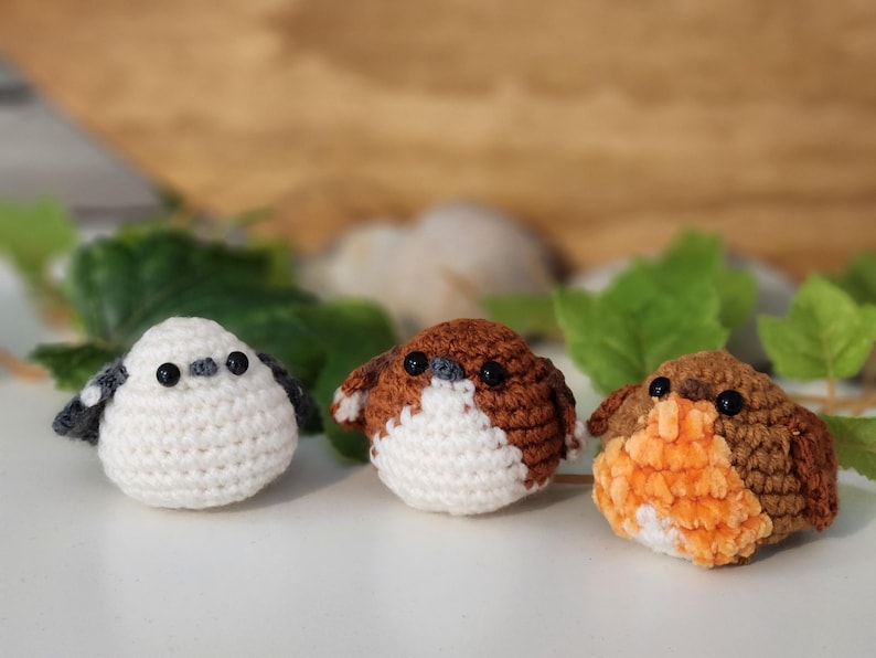 Puede incluir: Tres figuras de p&aacute;jaros de ganchillo hechas a mano. Los p&aacute;jaros son blancos, marrones y naranjas, con ojos negros. Son peque&ntilde;os y redondos, y parecen estar sobre una superficie blanca. Hay hojas verdes en el fondo.