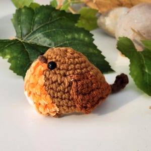 Puede incluir: Un petirrojo de ganchillo hecho a mano, con plumaje marr&oacute;n y naranja, un ojo negro y una peque&ntilde;a cola. El p&aacute;jaro se encuentra sobre un fondo de hojas verdes y una superficie clara, creando una est&eacute;tica natural y r&uacute;stica.