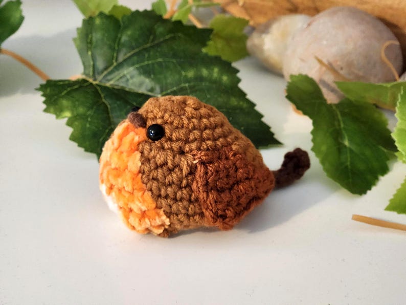 Puede incluir: Un petirrojo de ganchillo hecho a mano, con plumaje marr&oacute;n y naranja, un ojo negro y una peque&ntilde;a cola. El p&aacute;jaro se encuentra sobre un fondo de hojas verdes y una superficie clara, creando una est&eacute;tica natural y r&uacute;stica.
