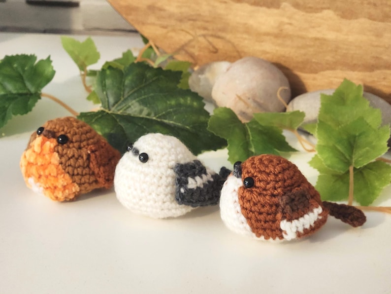 Puede incluir: Tres figuras de p&aacute;jaros de ganchillo en tonos marr&oacute;n, blanco y naranja. Los p&aacute;jaros est&aacute;n dispuestos en fila, con hojas verdes y peque&ntilde;as piedras en el fondo. Los p&aacute;jaros tienen ojos de cuentas negras y est&aacute;n hechos a mano.
