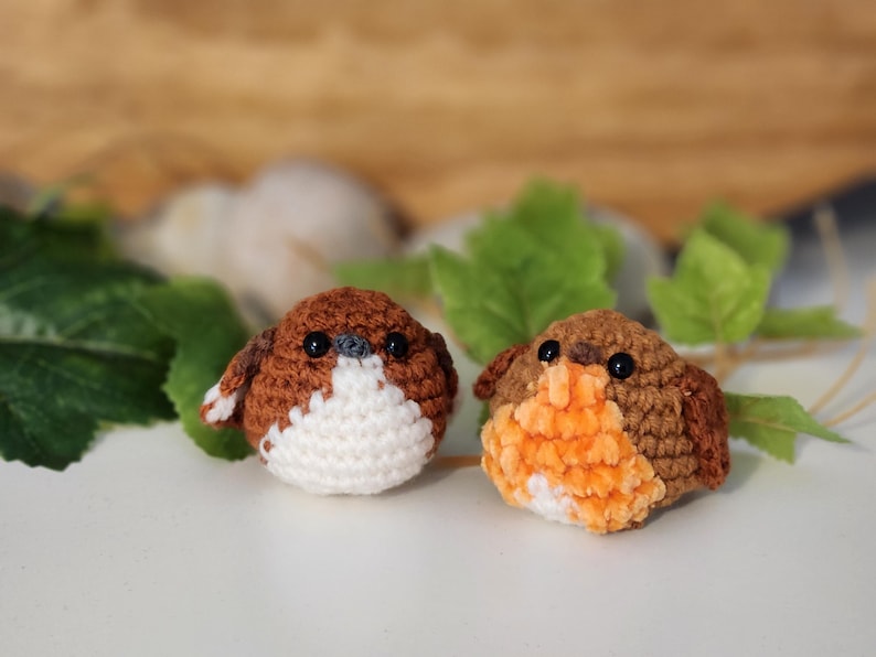 Puede incluir: Dos p&aacute;jaros de ganchillo hechos a mano. Uno tiene el pecho blanco y el otro naranja. Ambos p&aacute;jaros tienen alas y espalda marrones, con ojos negros. Los p&aacute;jaros est&aacute;n sobre una superficie blanca con hojas verdes en el fondo.
