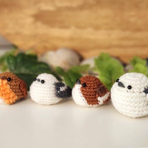 Puede incluir: Cuatro figuras de p&aacute;jaros de ganchillo hechas a mano en varios colores. Los p&aacute;jaros son blancos, marrones y naranjas, con ojos de cuentas negras. Est&aacute;n dispuestos en fila sobre una superficie blanca, con hojas verdes y un fondo de madera.