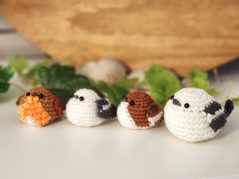 Puede incluir: Cuatro figuras de p&aacute;jaros de ganchillo hechas a mano en varios colores. Los p&aacute;jaros son blancos, marrones y naranjas, con ojos de cuentas negras. Est&aacute;n dispuestos en fila sobre una superficie blanca, con hojas verdes y un fondo de madera.