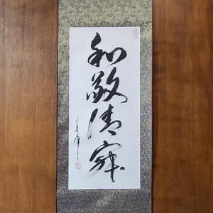 Könnte beinhalten: Eine traditionelle japanische Kalligraphie-Rolle mit schwarzen Tinte-Zeichen auf weißem Papier. Die Rolle hat einen gold-grauen gemusterten Rand und einen roten Siegelstempel. Die Kalligraphie ist auf einem Holzrahmen montiert.