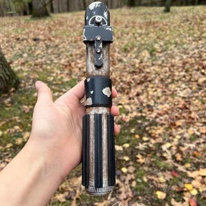 Puede incluir: Una r&eacute;plica de sable de luz de utiler&iacute;a de color negro y plateado desgastado con un acabado desgastado y descolorido. El sable de luz se sostiene en una mano, con la hoja retirada.