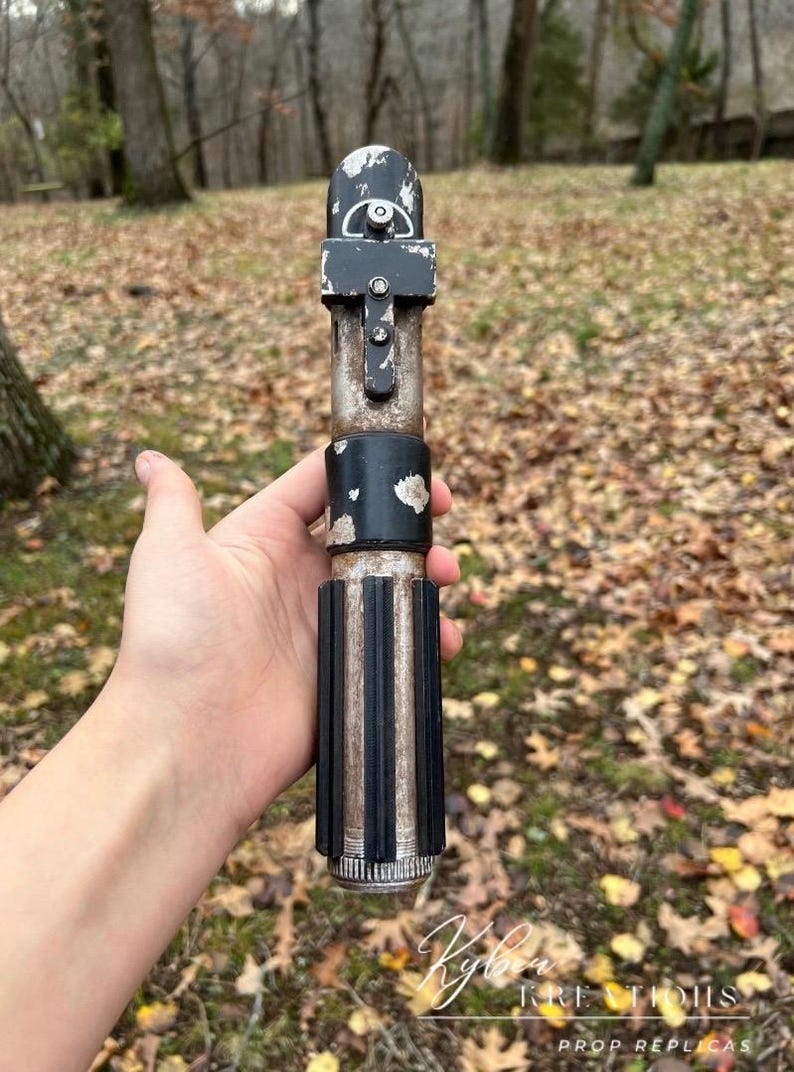 Puede incluir: Una r&eacute;plica de sable de luz de utiler&iacute;a de color negro y plateado desgastado con un acabado desgastado y descolorido. El sable de luz se sostiene en una mano, con la hoja retirada.