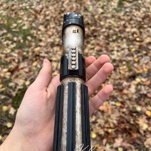 Puede incluir: Una r&eacute;plica de sable de luz de propulsi&oacute;n de plata y negro desgastada con un agarre negro y una hoja de plata. El sable de luz tiene una banda negra con una fila de puntos plateados. El texto "Kyber Kreations Prop Replicas" es visible en la parte inferior de la imagen.