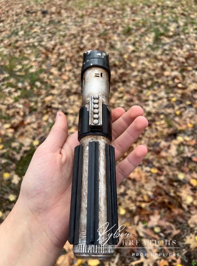Puede incluir: Una r&eacute;plica de sable de luz de propulsi&oacute;n de plata y negro desgastada con un agarre negro y una hoja de plata. El sable de luz tiene una banda negra con una fila de puntos plateados. El texto "Kyber Kreations Prop Replicas" es visible en la parte inferior de la imagen.