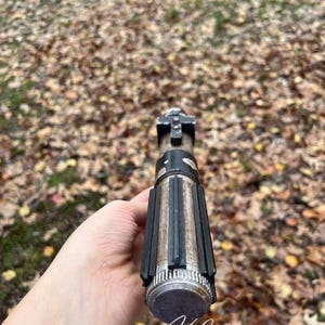 Puede incluir: Una empu&ntilde;adura de sable de luz desgastada, negra y plateada, con una superficie texturizada y un agarre negro. La empu&ntilde;adura se sostiene en una mano, con la punta de la empu&ntilde;adura mirando hacia la c&aacute;mara.