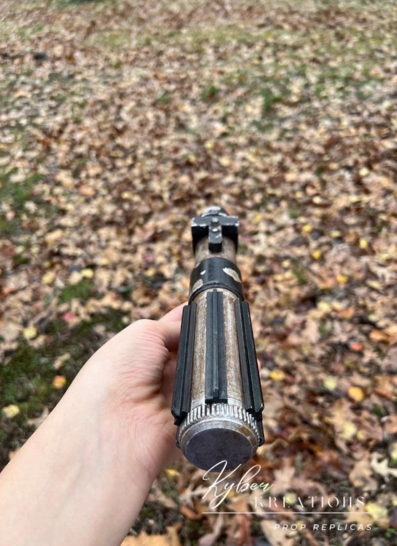 Puede incluir: Una empu&ntilde;adura de sable de luz desgastada, negra y plateada, con una superficie texturizada y un agarre negro. La empu&ntilde;adura se sostiene en una mano, con la punta de la empu&ntilde;adura mirando hacia la c&aacute;mara.