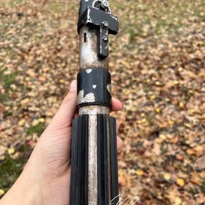 Puede incluir: Una r&eacute;plica de sable de luz de pl&aacute;stico, de color negro y plateado, con un mango detallado y una empu&ntilde;adura negra. El sable de luz se sostiene en una mano, con un fondo borroso de hojas ca&iacute;das.