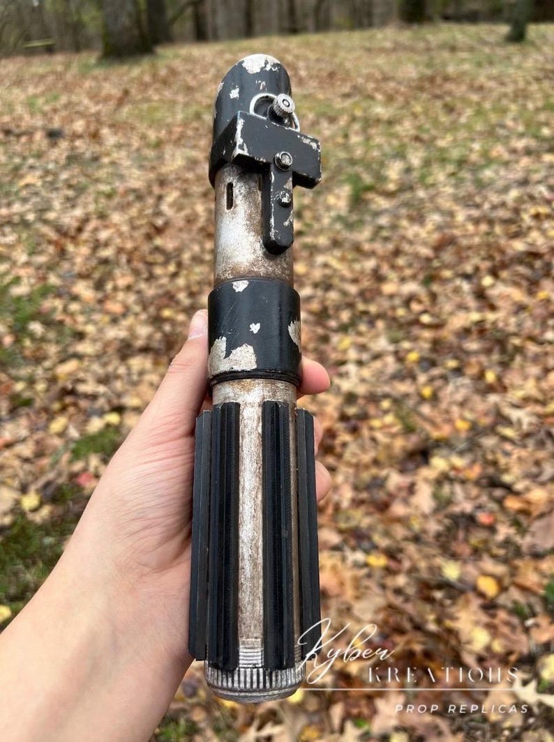 Puede incluir: Una r&eacute;plica de sable de luz de pl&aacute;stico, de color negro y plateado, con un mango detallado y una empu&ntilde;adura negra. El sable de luz se sostiene en una mano, con un fondo borroso de hojas ca&iacute;das.