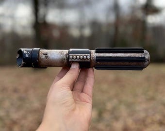 Könnte beinhalten: Eine verwitterte schwarze und silberne Lichtschwert-Requisite mit einer abgenutzten Metalloberfläche und einem schwarzen Griff. Das Lichtschwert wird in einer Hand vor einem verschwommenen Hintergrund aus Bäumen und Blättern gehalten.