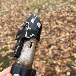 Puede incluir: Primer plano de una empu&ntilde;adura de sable l&aacute;ser negra y plateada con un acabado desgastado. La empu&ntilde;adura tiene un peque&ntilde;o pomo plateado en la parte superior y una correa negra alrededor del medio. La empu&ntilde;adura est&aacute; parcialmente oculta por una mano que la sostiene.