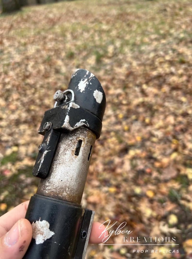 Puede incluir: Primer plano de una empu&ntilde;adura de sable l&aacute;ser negra y plateada con un acabado desgastado. La empu&ntilde;adura tiene un peque&ntilde;o pomo plateado en la parte superior y una correa negra alrededor del medio. La empu&ntilde;adura est&aacute; parcialmente oculta por una mano que la sostiene.