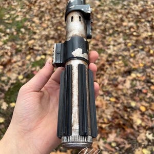 Puede incluir: Una r&eacute;plica de sable de luz plateado y desgastado con un mango negro y una hoja plateada desgastada. El sable de luz se sostiene en una mano, con el fondo desenfocado.