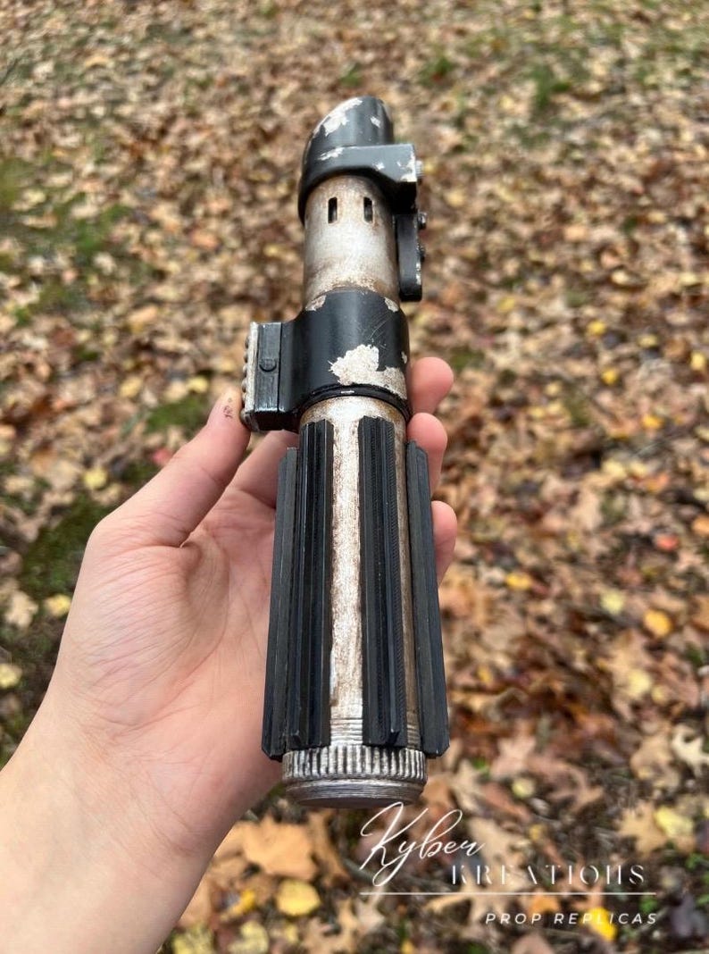 Puede incluir: Una r&eacute;plica de sable de luz plateado y desgastado con un mango negro y una hoja plateada desgastada. El sable de luz se sostiene en una mano, con el fondo desenfocado.