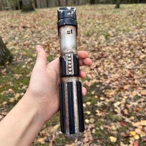 Puede incluir: Una r&eacute;plica de sable de luz de pl&aacute;stico, de color negro y plateado, con un acabado desgastado y envejecido. El sable de luz tiene un mango negro con una banda plateada y una hoja negra. El sable de luz se sostiene en una mano sobre un fondo de hojas secas.