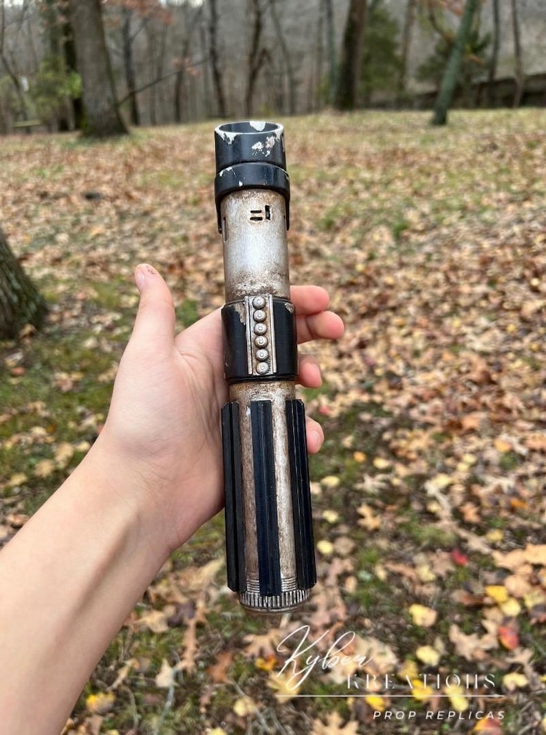 Puede incluir: Una r&eacute;plica de sable de luz de pl&aacute;stico, de color negro y plateado, con un acabado desgastado y envejecido. El sable de luz tiene un mango negro con una banda plateada y una hoja negra. El sable de luz se sostiene en una mano sobre un fondo de hojas secas.