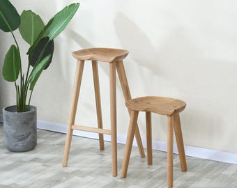 May include: Two wooden stools with a saddle-shaped seat. The taller stool is a bar stool with a simple design. The shorter stool is a smaller version of the bar stool.