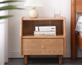 May include: A light wood nightstand with a single drawer and an open shelf. The nightstand has a small brass knob on the drawer and sits on four short legs. A lamp with a white pleated shade and a white ceramic base sits on the top shelf. A glass of water sits on the top shelf.
