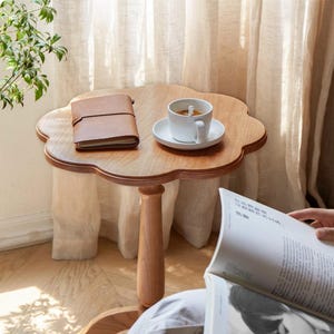 May include: A wooden side table with a flower-shaped top. The table has a single, turned leg and is decorated with a brown leather journal and a white coffee cup on a saucer.