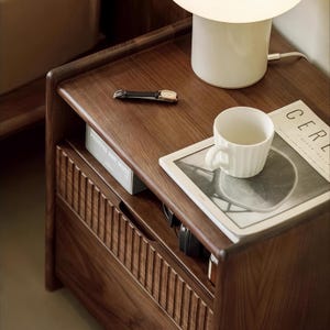 Puede incluir: Una mesita de noche de madera con un caj&oacute;n y un estante abierto. La mesita de noche tiene un acabado marr&oacute;n oscuro y est&aacute; decorada con una l&aacute;mpara de cer&aacute;mica blanca, una taza de cer&aacute;mica blanca, una revista y un reloj.