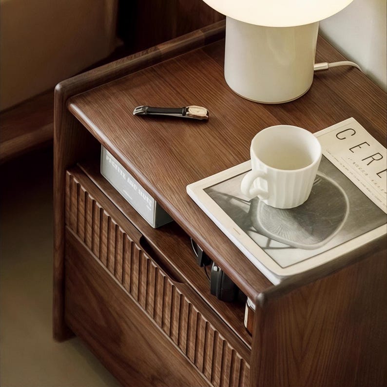 Puede incluir: Una mesita de noche de madera con un caj&oacute;n y un estante abierto. La mesita de noche tiene un acabado marr&oacute;n oscuro y est&aacute; decorada con una l&aacute;mpara de cer&aacute;mica blanca, una taza de cer&aacute;mica blanca, una revista y un reloj.