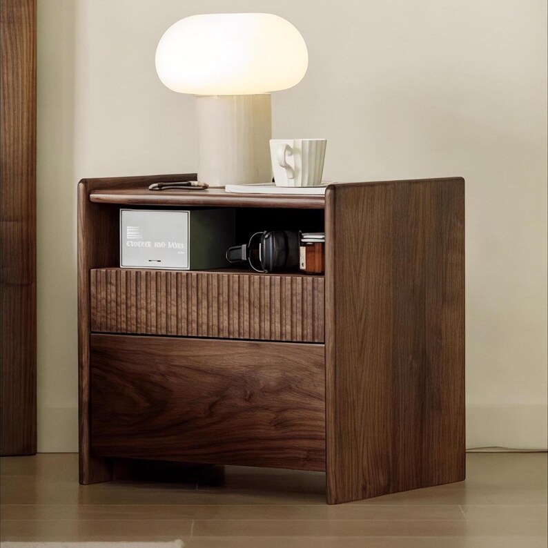 Puede incluir: Una mesita de noche de madera marr&oacute;n oscuro con dos cajones y un estante abierto. La mesita de noche tiene una l&aacute;mpara con una pantalla blanca y una taza blanca en la parte superior.