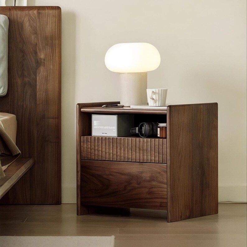 Puede incluir: Una mesita de noche de madera oscura con dos cajones y un estante abierto. La mesita de noche tiene una l&aacute;mpara blanca con una pantalla en forma de hongo en la parte superior. Hay una taza y un libro peque&ntilde;o en el estante.