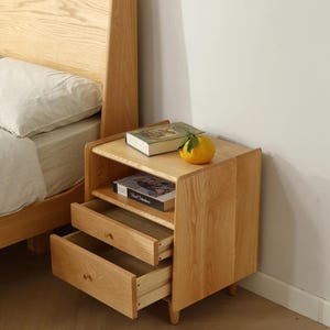 May include: A light wood bedside table with two drawers and an open shelf. The table has a natural wood finish and is accented with brass drawer pulls. A book and a citrus fruit are on the top shelf.