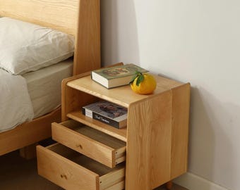 May include: A light wood bedside table with two drawers and an open shelf. The table has a natural wood finish and is accented with brass drawer pulls. A book and a citrus fruit are on the top shelf.