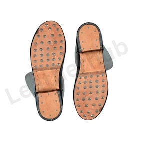 May include: Two black leather boots with visible soles. The soles are light brown with numerous metal studs. The boots have a simple design, with a focus on durability and functionality. The boots are likely for work or historical reenactment.
