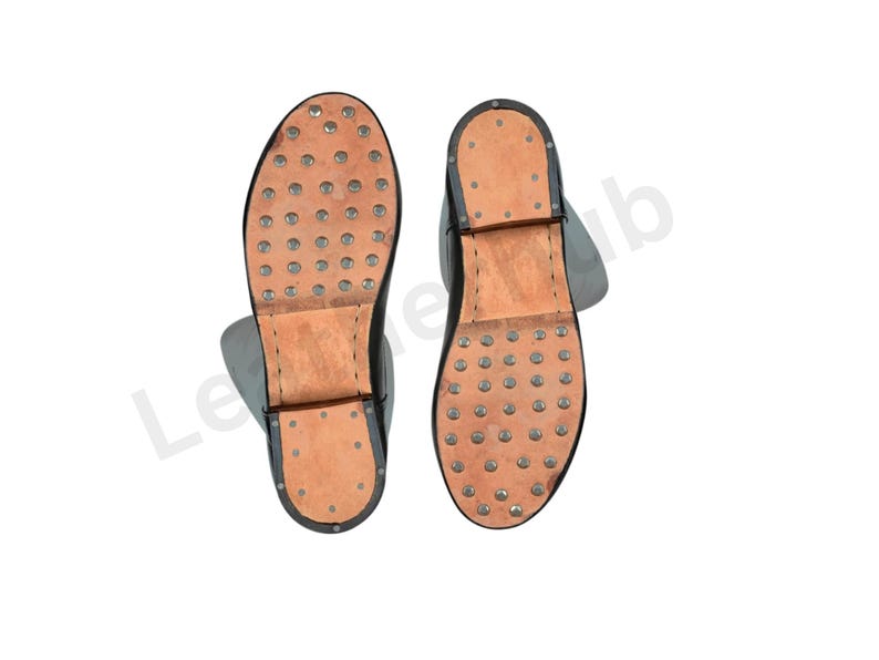 May include: Two black leather boots with visible soles. The soles are light brown with numerous metal studs. The boots have a simple design, with a focus on durability and functionality. The boots are likely for work or historical reenactment.