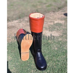 May include: A pair of tall black leather boots with a red cuff. One boot is upright, the other is lying on its side, revealing a light brown sole. The boots are on a grassy surface.