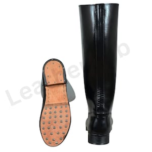 May include: Black leather riding boots. One boot is upright, showing the full length, while the other is lying on its side, displaying the sole with metal studs. The boots have a classic design with a smooth, shiny finish.