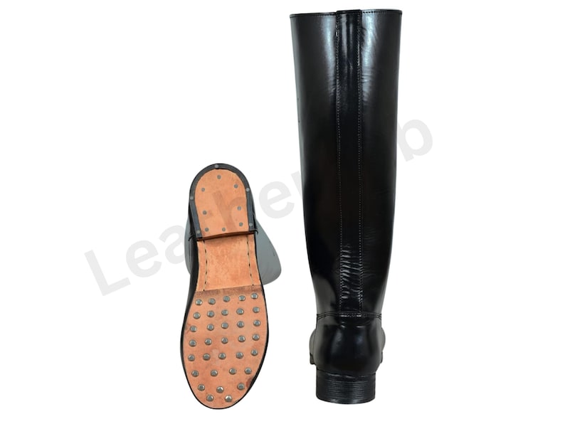 May include: Black leather riding boots. One boot is upright, showing the full length, while the other is lying on its side, displaying the sole with metal studs. The boots have a classic design with a smooth, shiny finish.