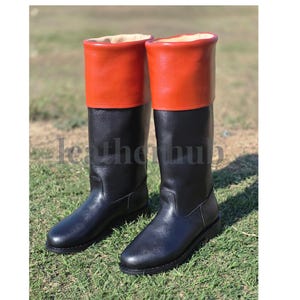 May include: A pair of tall black leather boots with orange tops, set against a backdrop of green grass. The boots have a classic design, suitable for various outdoor activities.