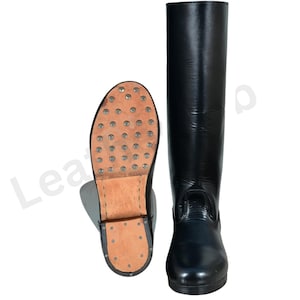 May include: Black leather riding boots. One boot is upright, the other is lying on its side, showing the sole with metal studs. The boots are tall, reaching the knee, and have a smooth, shiny finish. The soles are made of brown leather.