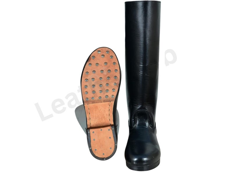 May include: Black leather riding boots. One boot is upright, the other is lying on its side, showing the sole with metal studs. The boots are tall, reaching the knee, and have a smooth, shiny finish. The soles are made of brown leather.