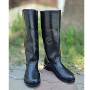 May include: A pair of black knee-high boots with a smooth finish, featuring a structured shaft, a rounded toe, and a low heel. The boots have a clean, minimalist design with vertical seam detailing along the front and sides of the leg.