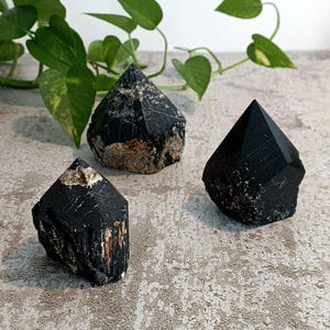 Könnte beinhalten: Drei schwarze Turmalin-Kristallspitzen, jede mit einer einzigartigen Form und Textur. Die Kristalle sind auf einer grauen Oberfläche angeordnet.