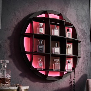 May include: A circular, dark wooden display shelf with several compartments, lit with a pink light. The shelf holds various perfume bottles. A decorative table and a pink chair are in the background.