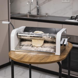 May include: A white and silver countertop pasta roller machine with a handle on the left side. The machine is rolling out a piece of dough, and there is flour on the surface of the machine.