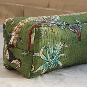 May include: Green quilted cosmetic bag with a jungle-themed print featuring a leopard, plants, and trees. The bag has a rectangular shape with a zipper closure and red trim. The bag is made of a soft, quilted fabric.
