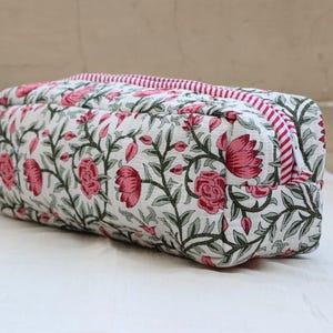 May include: A rectangular, quilted cosmetic bag with a floral pattern. The bag features a white background with pink roses and green leaves. The zipper and trim are striped in pink and white.