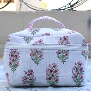 Può includere: Una borsa cosmetica trapuntata rettangolare in tessuto bianco con un motivo floreale ripetuto di petali rosa e foglie verdi. La borsa include un manico superiore con un design a righe rosa e bianche e una chiusura a zip bianca sul bordo.
