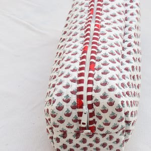 May include: A cylindrical pencil case against a white backdrop. The case features a repeating pattern of maroon and teal floral designs. A red and white striped border surrounds the open zipper.