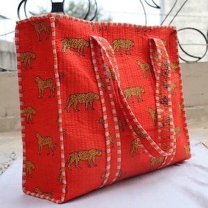 May include: A large, rectangular tote bag in a vibrant orange hue, decorated with a repeating pattern of tan leopards. The bag has sturdy handles and a striped border. The bag is made of a quilted fabric.