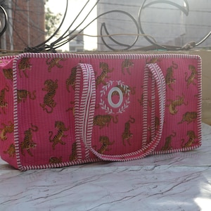 Puede incluir: Un bolso rectangular acolchado de color rosa con un patr&oacute;n repetitivo de tigres en tonos marrones y negros. El bolso tiene bordes y asas a rayas blancas y rosas, con una letra O bordada en blanco rodeada por un dise&ntilde;o de corona de laurel.